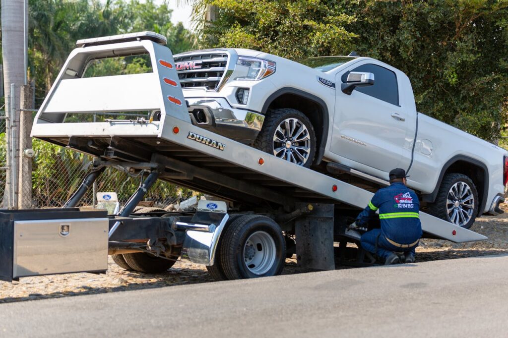 Tow truck operator loading white GMC pickup truck on street in daytime.